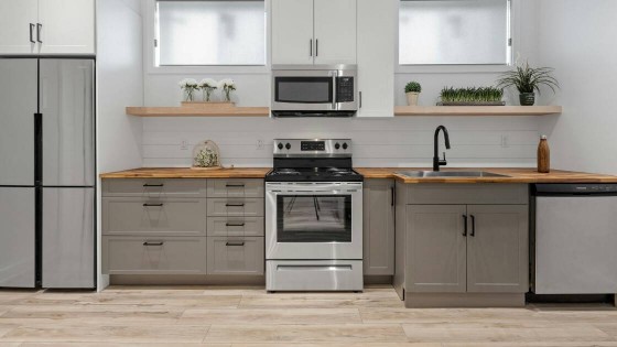 A modern kitchen with light gray lower cabinets, open wood shelves, and light wood flooring features a stainless steel refrigerator, microwave, range, and an under-counter dishwasher with its