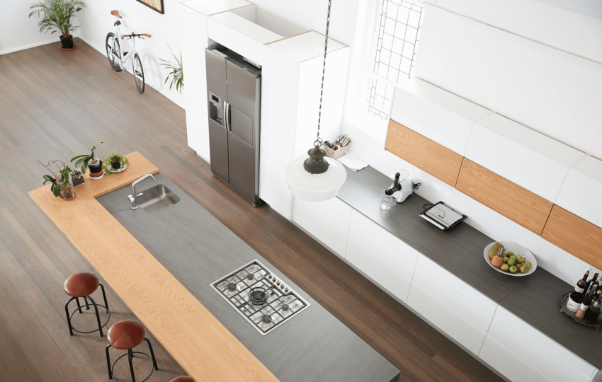 Spacious modern kitchen with a long island counter featuring a sink and stovetop. Bar stools, potted plants, a sleek fridge, and a bicycle are visible.