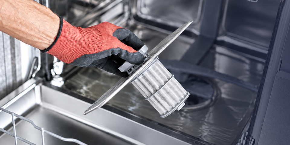 Close-up of a gloved hand lifting a dishwasher filter assembly out of the dishwasher for cleaning or inspection.